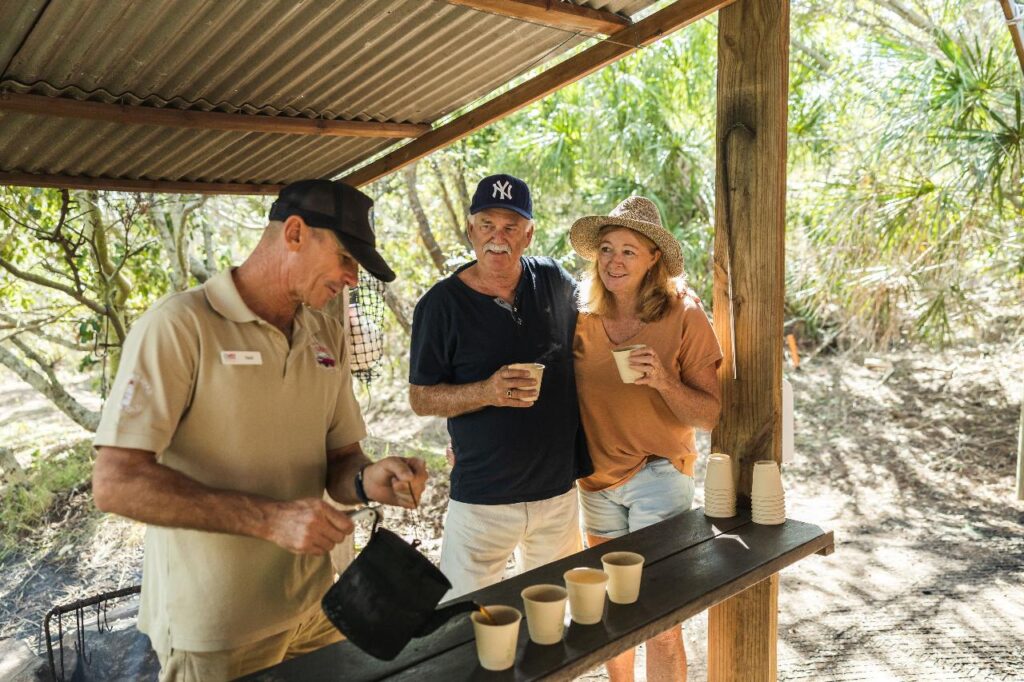 Three adults stand at an outdoor wooden counter as one pours drinks into paper cups; two others smile and hold cups, surrounded by greenery.
