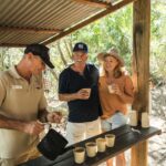 Three adults stand at an outdoor wooden counter as one pours drinks into paper cups; two others smile and hold cups, surrounded by greenery.