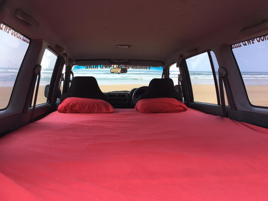 View from inside a vehicle with a red-covered mattress and pillows in the back, facing the beach and ocean through the open rear doors.