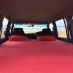 View from inside a vehicle with a red-covered mattress and pillows in the back, facing the beach and ocean through the open rear doors.