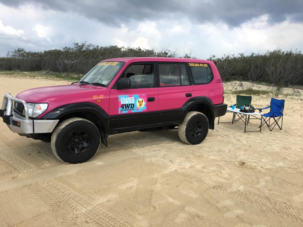 A pink 4WD vehicle is parked on sandy ground near two blue camping chairs and a small table with items, with bushes and cloudy sky in the background.