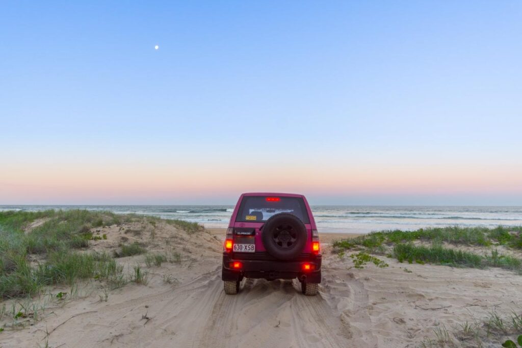 A red SUV parked on sandy dunes faces the ocean under a clear sky with a visible moon; grassy vegetation borders the scene.