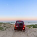 A red SUV parked on sandy dunes faces the ocean under a clear sky with a visible moon; grassy vegetation borders the scene.