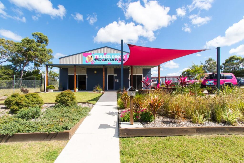A single-story building with a sign reading “Fraser Dinghi Adventures,” landscaped garden beds, and a red shade sail over the entrance on a sunny day.