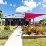 A single-story building with a sign reading “Fraser Dinghi Adventures,” landscaped garden beds, and a red shade sail over the entrance on a sunny day.