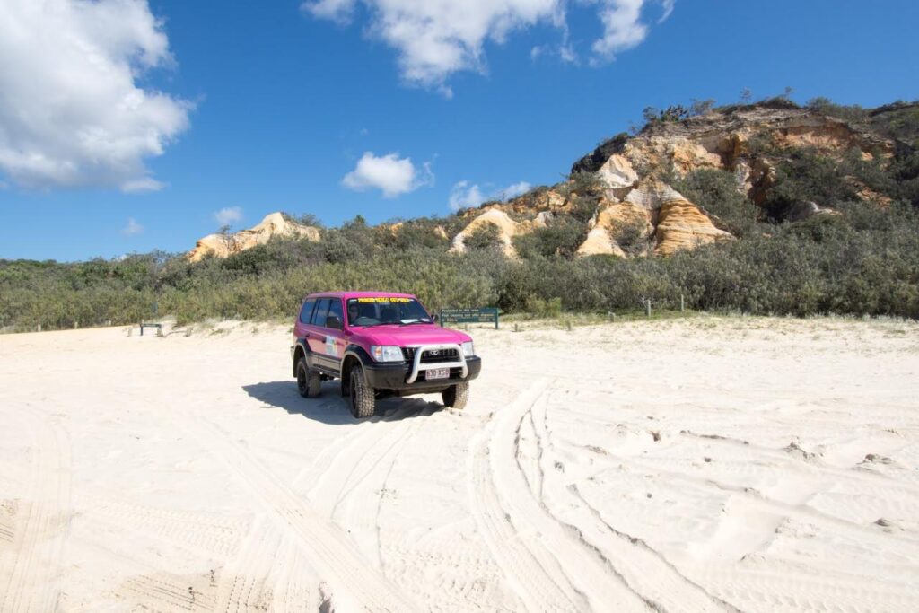 A pink SUV is parked on a sandy beach with tire tracks nearby, surrounded by bushes and rocky hills under a blue sky with clouds.