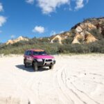 A pink SUV is parked on a sandy beach with tire tracks nearby, surrounded by bushes and rocky hills under a blue sky with clouds.