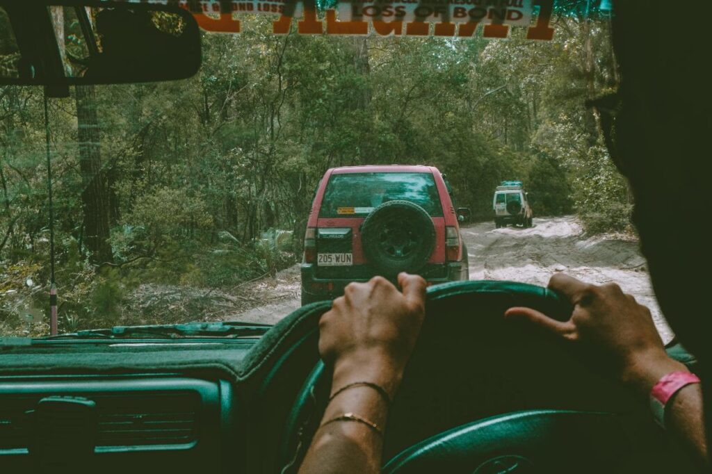 A person drives a car on a dirt road through a forest, following two other vehicles visible ahead.