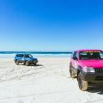 Two SUVs, one blue and one pink, are parked on a sandy beach under a clear blue sky, with the ocean visible in the background.