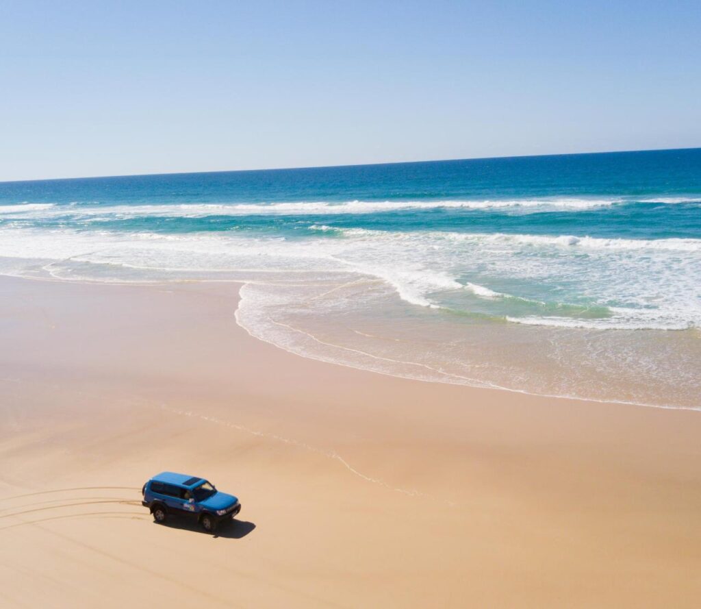 A blue SUV drives along an empty sandy beach near the shoreline with ocean waves under a clear blue sky.