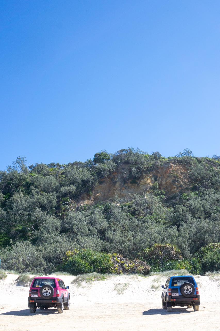 Two off-road vehicles are parked on sandy ground in front of a green hillside under a clear blue sky.