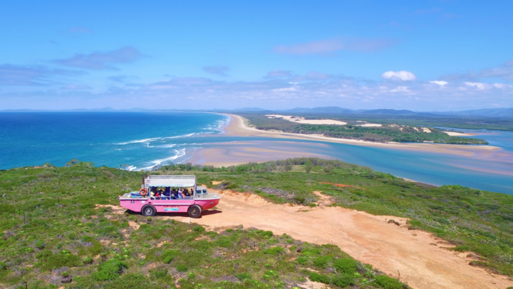 A pink amphibious tour vehicle drives along a coastal dirt path with a scenic view of the beach, ocean, sand dunes, and greenery under a clear blue sky.