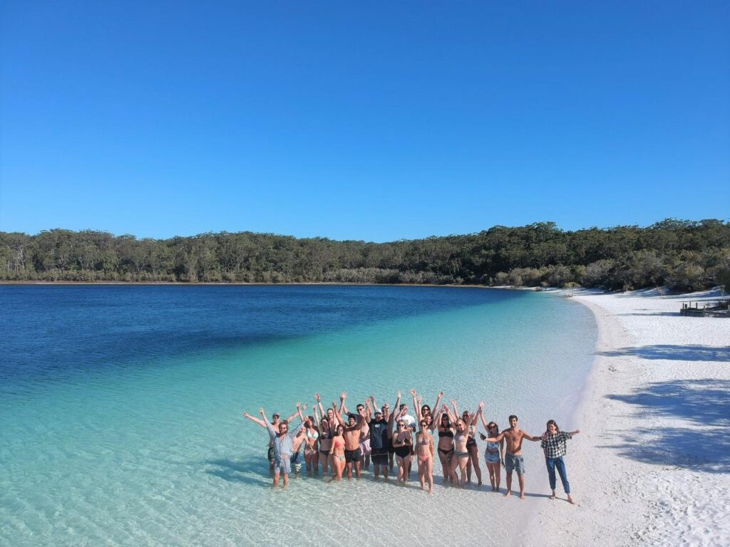 A group of people stand in shallow water on a white sand beach, raising their arms, with turquoise water and a forested shoreline under a clear blue sky.