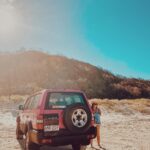 A person stands next to a red SUV parked on sandy terrain with hills and trees in the background under a bright, sunny sky.