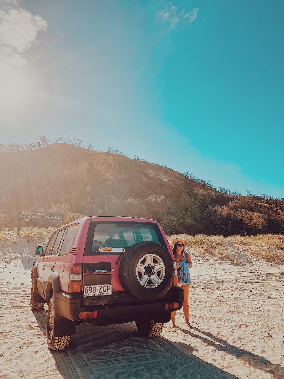A person stands next to a red SUV parked on sandy terrain with hills and trees in the background under a bright, sunny sky.
