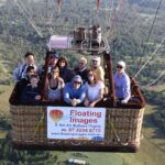 A group of people in a hot air balloon basket with a "Floating Images Hot Air Balloon Flights" sign, flying over a green landscape.