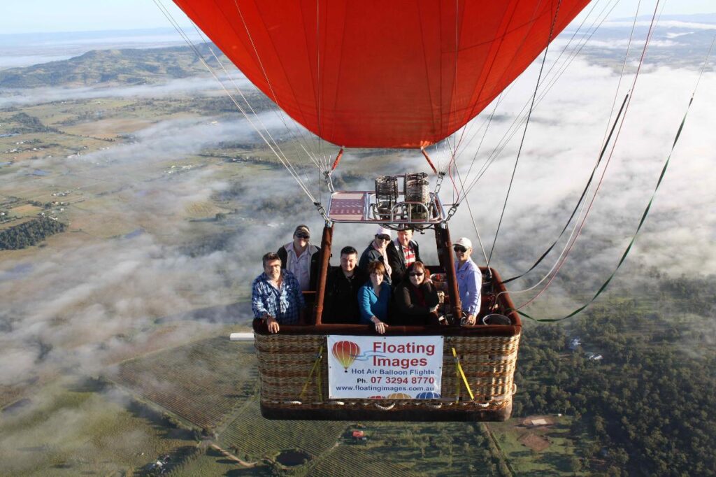A group of people ride in a hot air balloon basket high above a rural landscape with fog and farmlands visible below.