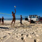 A group of people play beach volleyball on sand near the ocean, with a volleyball net and a parked white SUV in the background under a clear blue sky.