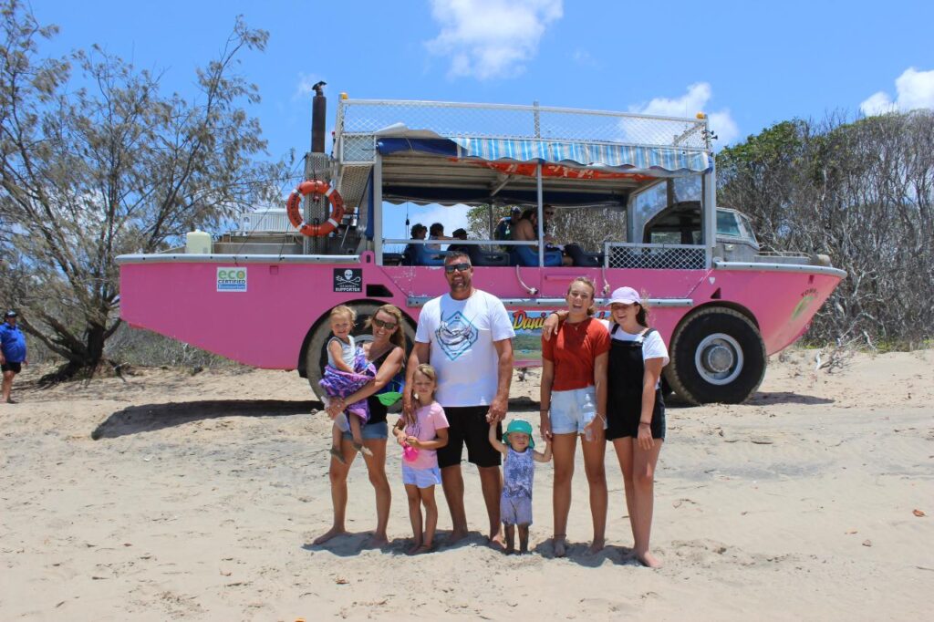 A group of people stand on a sandy beach in front of a pink amphibious tour vehicle with trees and blue sky in the background.
