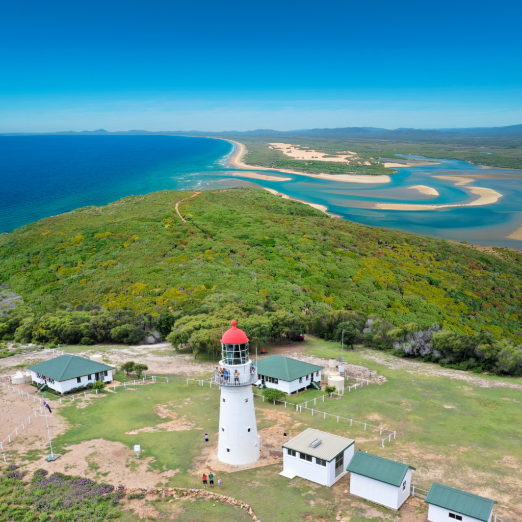 Aerial view of a white lighthouse with a red roof surrounded by small buildings, green hills, and coastline meeting the ocean and river inlets.