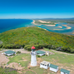 Aerial view of a white lighthouse with a red roof surrounded by small buildings, green hills, and coastline meeting the ocean and river inlets.