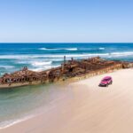A rusted shipwreck lies partially submerged at the edge of the ocean on a sandy beach, with a pink SUV parked nearby.