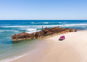 A rusted shipwreck lies partially submerged at the edge of the ocean on a sandy beach, with a pink SUV parked nearby.