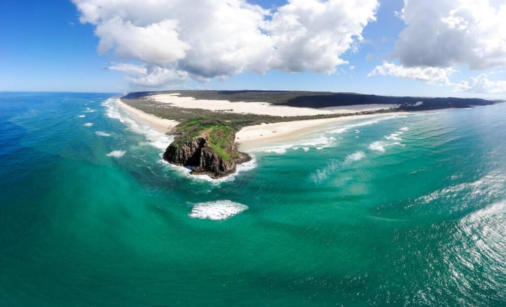 Aerial view of a sandy coastline with turquoise ocean waves, rocky headland, white sand dunes, and partly cloudy sky.