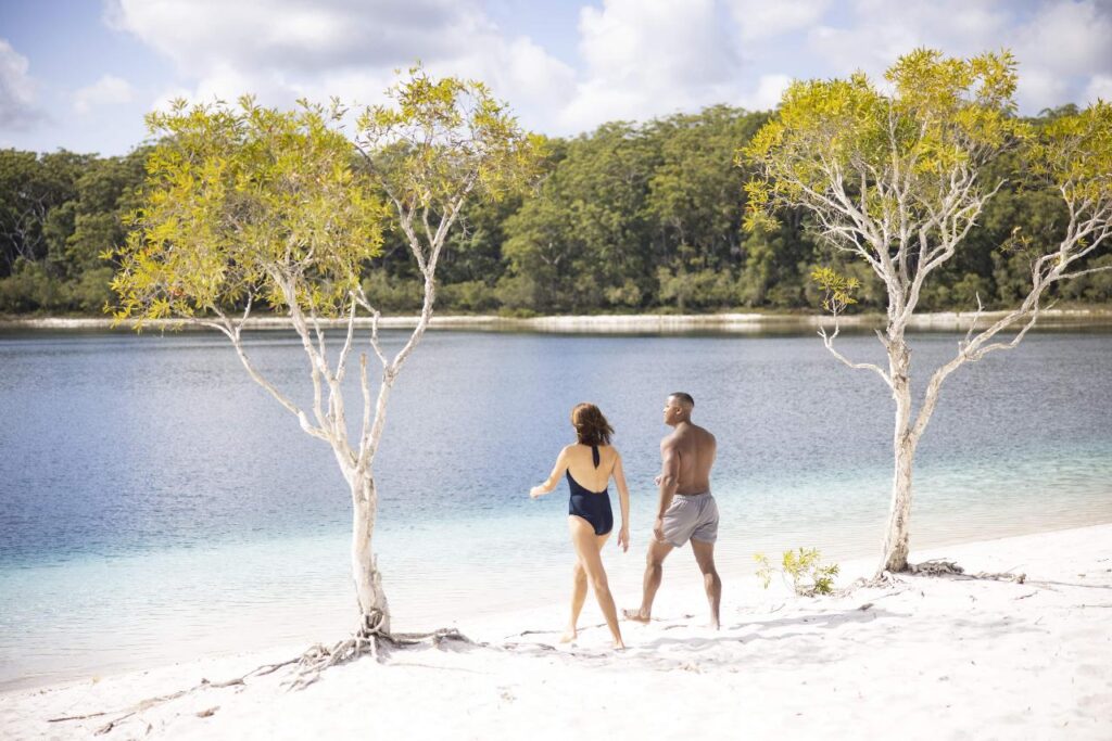 A woman and a man in swimsuits walk barefoot on white sand by a lake, with trees and forest in the background under a partly cloudy sky.