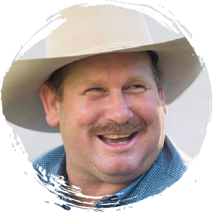 A man wearing a wide-brimmed hat and a blue plaid shirt smiles, with a trimmed mustache and light facial hair visible.