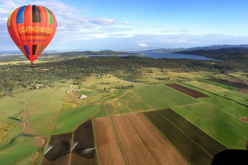 A colorful hot air balloon floats above patchwork farmland with fields, scattered houses, and a large lake in the background.