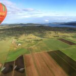 A colorful hot air balloon floats above patchwork farmland with fields, scattered houses, and a large lake in the background.