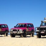 Three off-road vehicles, two pink and one white with a roof rack, are parked on a sandy beach under a clear blue sky.