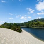 A sandy dune slopes down to a narrow, dark blue river bordered by dense green trees under a blue sky with scattered clouds.