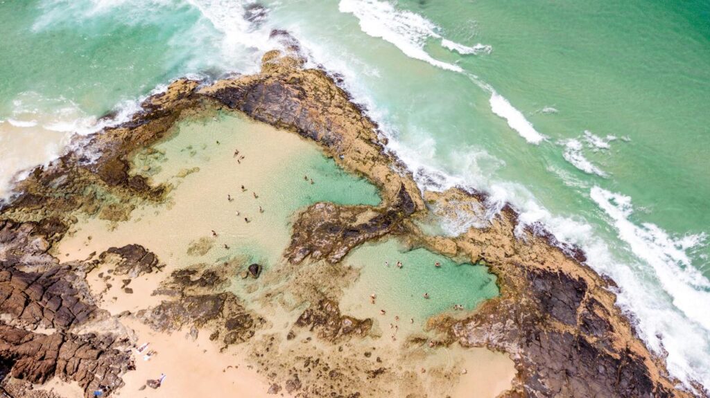 Aerial view of a natural tidal pool surrounded by rocks and sand at the edge of the ocean, with people swimming and relaxing in the water.