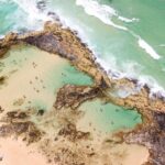 Aerial view of a natural tidal pool surrounded by rocks and sand at the edge of the ocean, with people swimming and relaxing in the water.