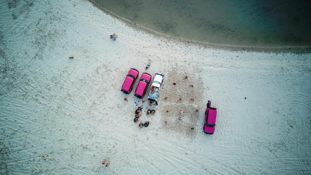 Aerial view of four vehicles parked on a sandy beach near water, with groups of people gathered around them.