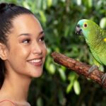 A woman smiles while looking at a green parrot perched on a branch, with green foliage in the background.