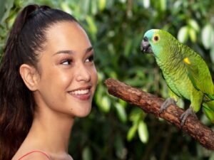 A woman smiles while looking at a green parrot perched on a branch, with green foliage in the background.
