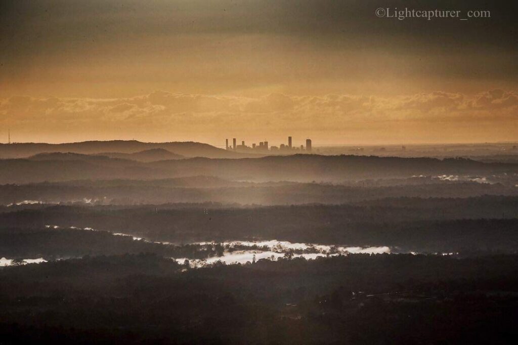A distant city skyline rises above layers of misty hills and reflective water under a cloudy, golden sky at sunrise or sunset.