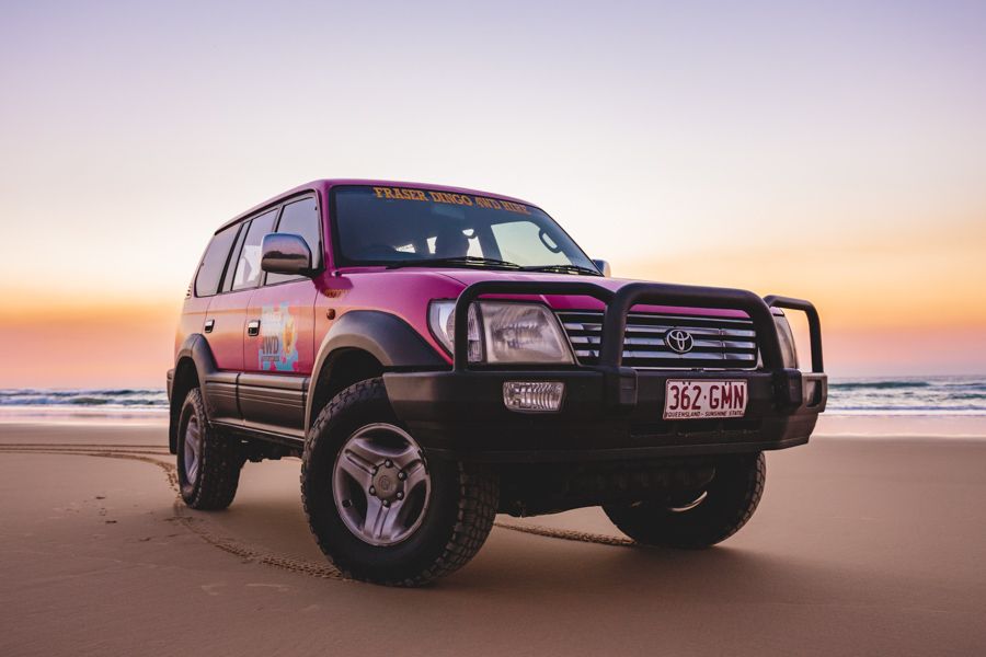 A red Toyota SUV with off-road tires is parked on a sandy beach at sunset, with the ocean and pastel sky in the background.