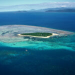 A small, lush island is surrounded by turquoise coral reefs and deep blue ocean, with distant islands visible on the horizon under a partly cloudy sky.