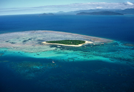 A small, lush island is surrounded by turquoise coral reefs and deep blue ocean, with distant islands visible on the horizon under a partly cloudy sky.