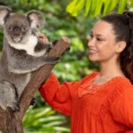 A woman in an orange top stands next to a koala perched on a tree branch, both set against a lush, green natural background.
