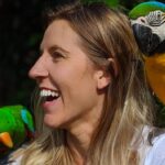 A woman with long blonde hair smiles while two colorful parrots perch on her shoulders, one green and one blue and yellow.
