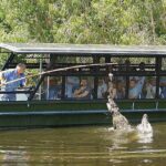 A tour guide on a boat uses a stick to interact with a jumping crocodile in the water, while passengers watch from behind protective bars.