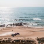 Aerial view of a beach with people walking near a large, rusted shipwreck partially submerged in the ocean and several vehicles parked on the sand.