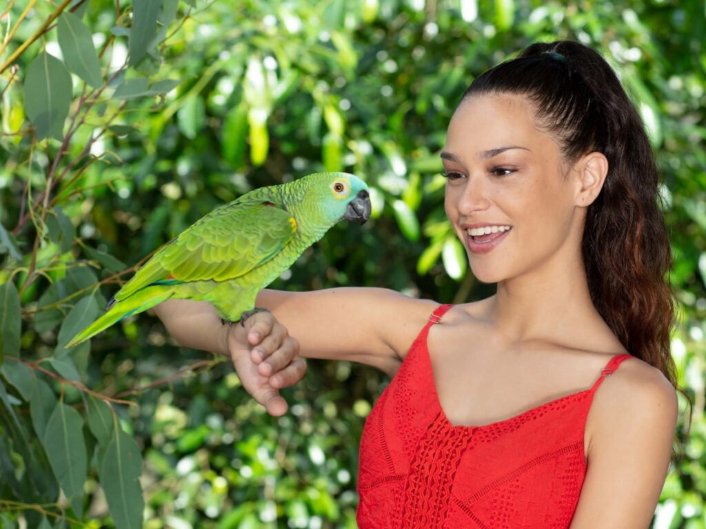 Woman in a red dress smiling at a green parrot perched on her arm, with green foliage in the background.