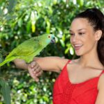 Woman in a red dress smiling at a green parrot perched on her arm, with green foliage in the background.