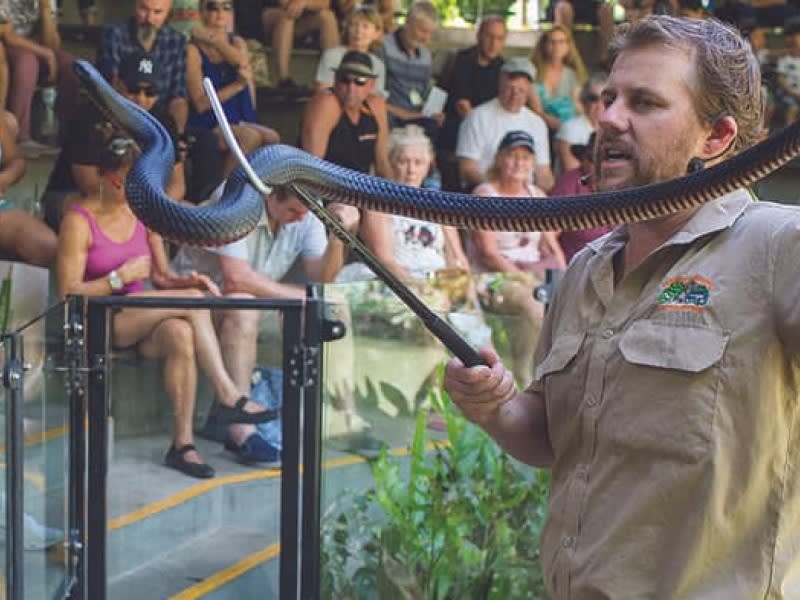 A man holding a large snake with a snake hook stands in front of an audience seated on bleachers.